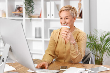 Wall Mural - Mature female programmer with cup of coffee working at table in office