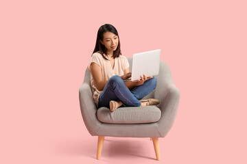 Wall Mural - Beautiful Asian woman using laptop in grey armchair on pink background