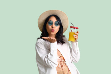 Wall Mural - Beautiful Asian woman with glass of juice blowing kiss on green background