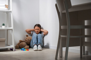 Stressed little Asian girl with autistic disorder and toy bear sitting in corner at home