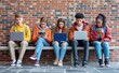 © ProstoSvet - Group of multicultural teenagers engaged with laptops and smartphones sitting outside on campus.