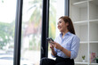 © Wasana - Smiling young Asian businesswoman using her smartphone by the window in a well-lit office, dressed in smart casual attire.
