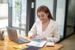 © Wasana - Professional Asian businesswoman typing on laptop at a wooden desk with paperwork and greenery in a bright office setting.