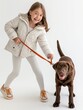 A joyful young girl in a white coat playing with her happy brown Labrador on a leash against a white background