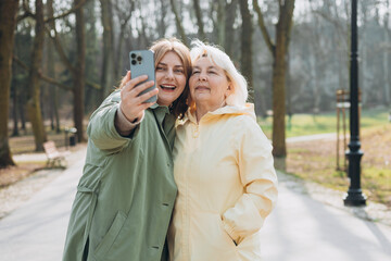 Wall Mural - Happy older Mother and adult positive daughter are doing Selfie on city street, Urban lifestyle concept. Travelers. Outdoor shot of well-dressed female friends. Family day