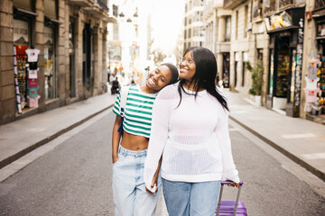 A young lesbian couple is exploring a new city during a short weekend trip, walking together while pulling a suitcase along a city street.