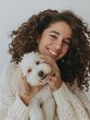A cheerful young woman with curly hair smiling while holding her cute white dog, conveying a warm moment of companionship.