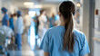 A nurse in scrubs. standing in a busy hospital ward with doctors and patients around. The focus is from behind her as she coordinates her teams work during visiting hours