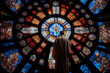 Beautiful artwork on a huge stained glass window towering over a priest, casting a solemn mood as he leads a religious congregation in prayer and mass during Sunday worship inside a Christian church.
