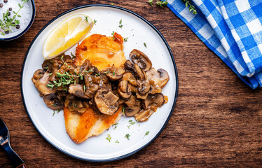 Wall Mural - French breaded chicken steak with mushrooms and onion sauce with white wine, lemon and thyme on plate, rustic table background, top view