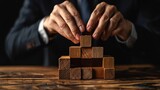 Fototapeta  - A business man attempts to construct a wooden block on a wooden table with a black backdrop as the foundation for a business organization.