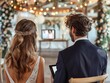 A bride and groom are sitting in a wedding ceremony. The bride is wearing a headband and the groom is wearing a suit. They are looking at a tablet or laptop