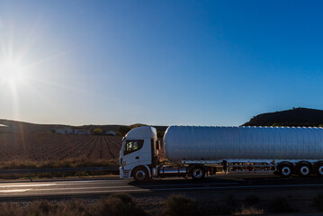 Canvas Print - Gas tanker truck driving on a conventional road with a sun at dawn, side view.