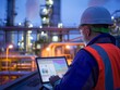Engineer in safety gear works on a laptop with industrial plant background.