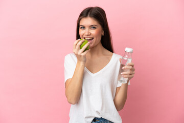 Wall Mural - Young caucasian woman isolated on pink background with a bottle of water and eating an apple