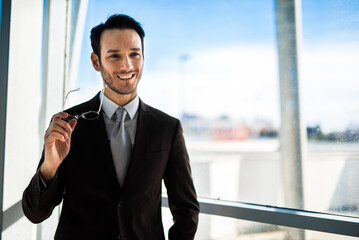 Smiling young man in a suit stands near a window with eyeglasses in hand