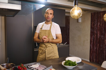 Wall Mural - Authentic portrait of male chef in beige kitchen apron, standing at kitchen counter with fresh ingredients, looking at camera, preparing dinner at home kitchen