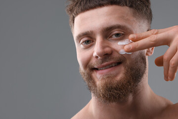 Poster - Handsome man applying moisturizing cream onto his face on grey background, space for text