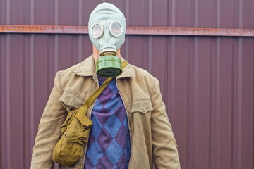 one military gray rubber protective gas mask with a green iron filter on the face of a man dressed in a dirty yellow jacket stands near a brown fence during the day on the street