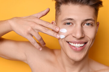 Canvas Print - Handsome man applying moisturizing cream onto his face on orange background, closeup