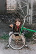 Little beautiful sad teenage girl, dissatisfied upset child showing dislike thumbs down sits near an old bicycle with a broken, punctured wheel tire outdoors. Photography, portrait.