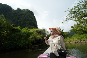 Wall Mural - A female model wearing a hijab is sitting on a wooden boat sailing around a river with many mangrove forests on its side