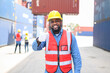 In container yards, African American container operators wear yellow helmets and safety vests look at the camera, and show thumbs up. Transportation import and export logistic industry concept.