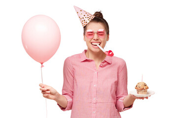 Young happy girl in glasses and birthday hat, blowing whistle, holding pink balloon and cupcake, laughing and enjoying party