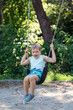 Smiling little boy swings hard holding chains against sunny summer nature background.