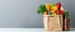 © Lasvu - Different grocery products in a paper bag placed on a white table against a gray wall. The bag contains a variety of fresh vegetables, fruits, pasta, and canned goods.