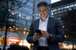 © Stock 4 You - Smiling mature hispanic senior business man using app, holding smartphone cellphone at office centre at night. Indian or latin middle age male entrepreneur businessman working on mobile cell phone.
