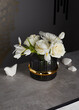 On a gray background, white flowers stand in a black vase, surrounded by stones.