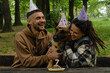 Dog celebrates birthday with family. Young couple hugs German Shepherd from both sides in spring green park. Dog birthday party with meat cake and 7 candle, paper hats on heads.