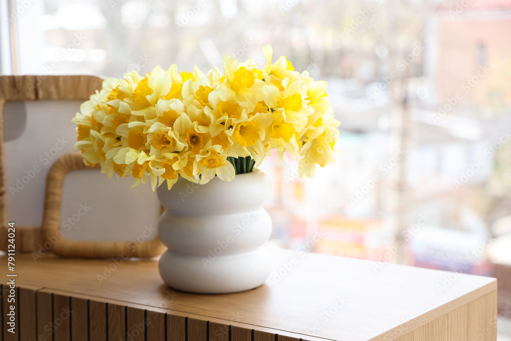 Vase with daffodils and frames on commode near window in room, closeup