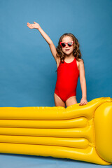 Swimming is fun. Happy little girl wearing red swim suit in sunglasses going to poll with yellow inflatable mattress, blue studio background. Summer vacation concept. Happy family