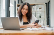 © Satori Studio - Smiling young woman in a white blazer sits at a wooden desk, working on a laptop and holding a smartphone.