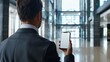 © rajagambar99 - photo shot of a businessman taken from his back, holding a modern smartphone showing a blank white screen, in a modern office, daytime, depth of field