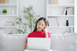 Happy young Asian woman wearing eyeglasses using laptop while seated on couch at home