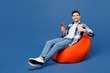 © ViDi Studio - Full body young man he wear shirt white t-shirt casual clothes sit in bag chair hold in hand use point finger on mobile cell phone isolated on plain blue background studio portrait. Lifestyle concept.