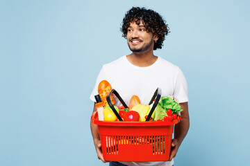 Wall Mural - Young Indian man in white t-shirt casual clothes hold red basket for takeaway mock up with food products look aside on area isolated on plain blue background Delivery service from shop or restaurant