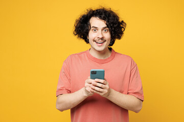 Young smiling excited fun happy man wear pink t-shirt casual clothes hold in hand use mobile cell phone chatting online isolated on plain yellow orange background studio portrait. Lifestyle concept