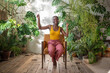 Carefree happy african american woman in headphones enjoying music among houseplants. Joyful smiling black girl sitting on chair at home tropical garden listening favourite track and lightly dancing.