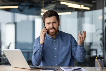 Wall Mural - Portrait of an angry young man businessman sitting at a desk and talking emotionally on a mobile phone, waving his hands