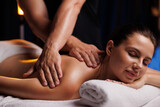 Fototapeta Łazienka - A young brunette woman receives a professional back massage in a spa salon. A beautiful naked lady in a towel with perfect skin gets a relaxing massage. The concept of luxury professional massage.