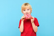 Cute little boy with blond hair and freckles eating a glazed donut. Boy holding colorful donuts, eating junk unhealthy food full of sugar, isolated on pastel blue studio background.