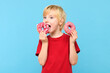 Cute little boy with blond hair and freckles eating a glazed donut. Boy holding colorful donuts, eating junk unhealthy food full of sugar, isolated on pastel blue studio background.