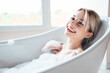 Portrait of young beautiful sexy woman having fun while lying in bathtub full of foam at home. Charming smiling model relaxing in luxury bath interior. Female enjoying beauty and skincare day