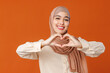 Beautiful muslim woman in hijab showing heart symbol on orange background