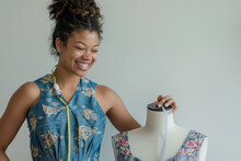 A Woman In A Blue Dress Taking Measurements Of The Dimple On Her Mannequin With A Tape Measure Around It And Smiling While Standing Near A White Wall In A Home Studio