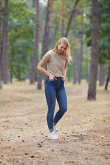 Wall Mural - Blue-eyed blonde in a beige T-shirt walks in a pine forest. Portrait of a joyful young woman enjoying in autumn park.